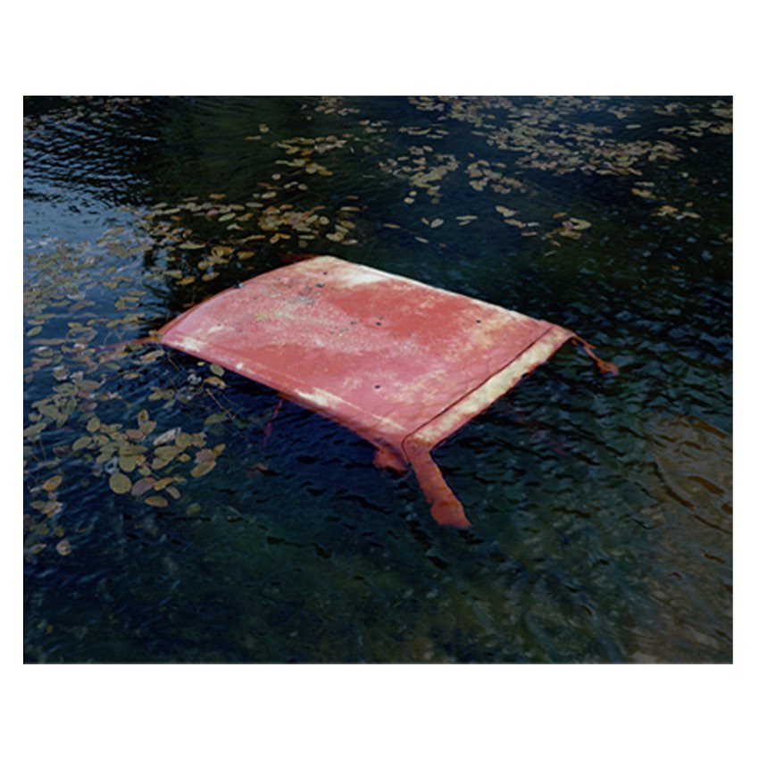 An old, rusty car roof partially submerged in dark water with lily pads scattered around, creating a surreal and abandoned scene.