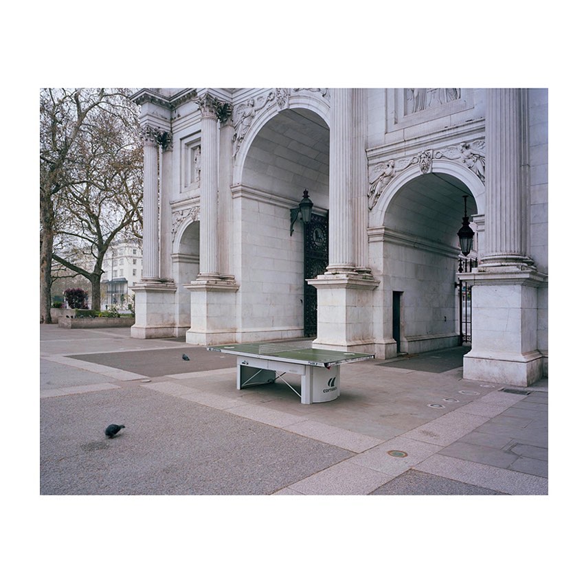 Outdoor shot of an empty ping pong table on a stone square with archways, columns, trees, and buildings in the background during daytime.