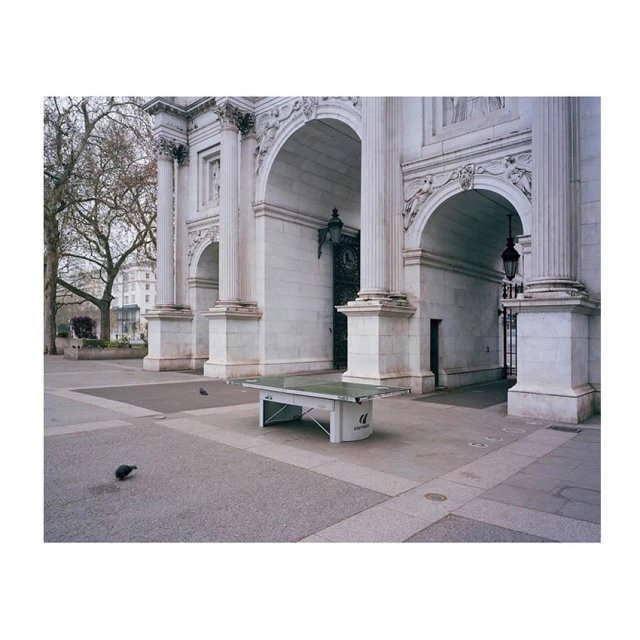 Outdoor shot of an empty ping pong table on a stone square with archways, columns, trees, and buildings in the background during daytime.