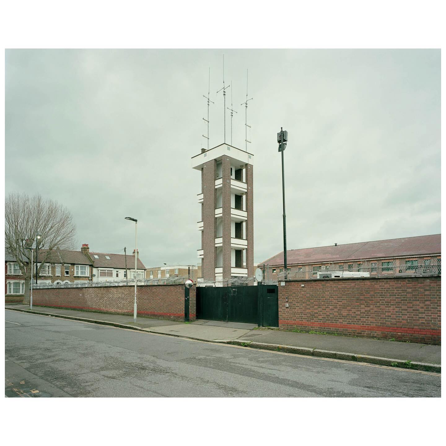 Tall brick tower with antennas on top stands near a brick wall and buildings under an overcast sky in an urban environment.