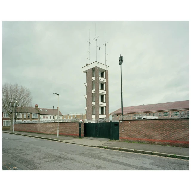 Tall brick tower with antennas on top stands near a brick wall and buildings under an overcast sky in an urban environment.