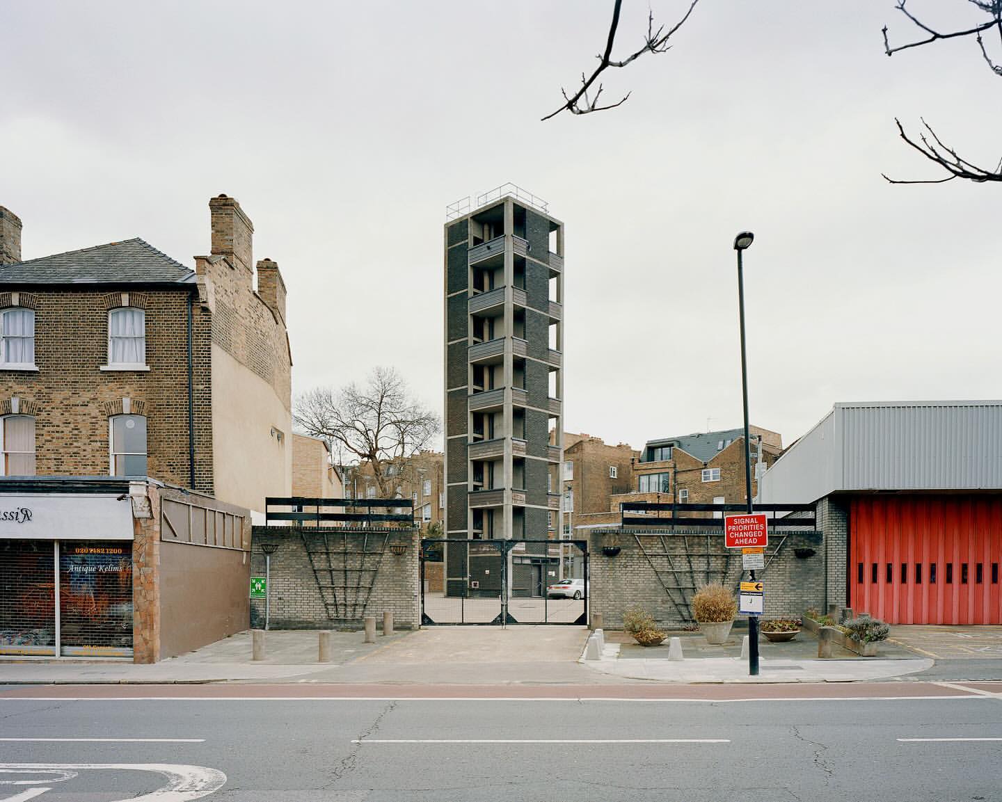 Exterior view of a tall, modern concrete building with residential balconies, set within an urban scene with brick buildings and a street.