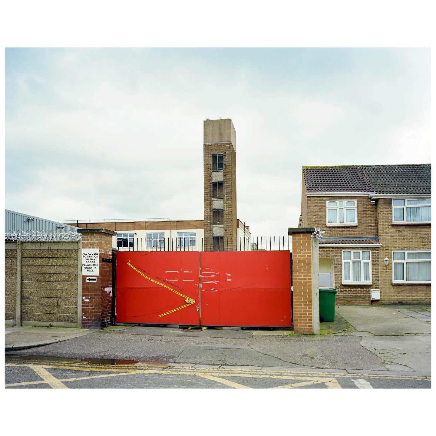 Wide outdoor shot of a large red metal gate dividing the street, with a tall brick building and houses in the background under cloudy sky.