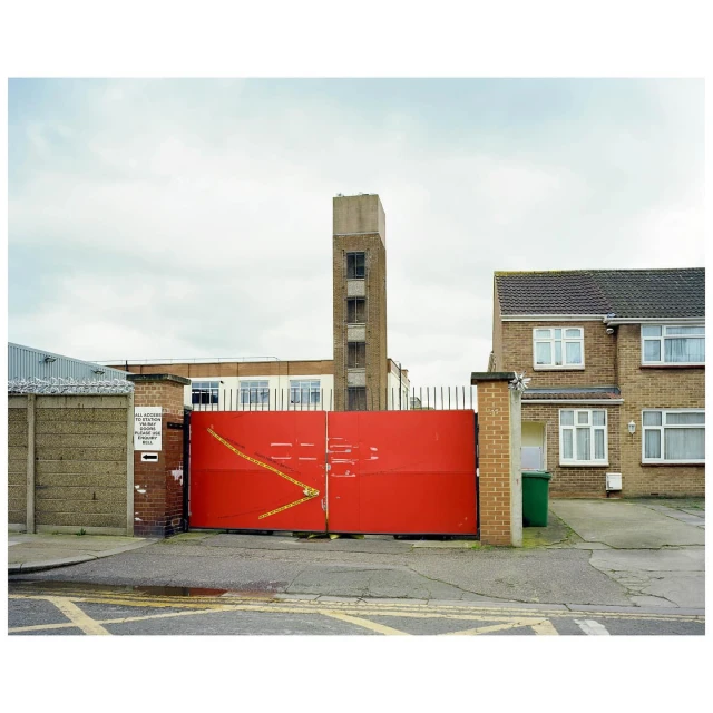 Wide outdoor shot of a large red metal gate dividing the street, with a tall brick building and houses in the background under cloudy sky.