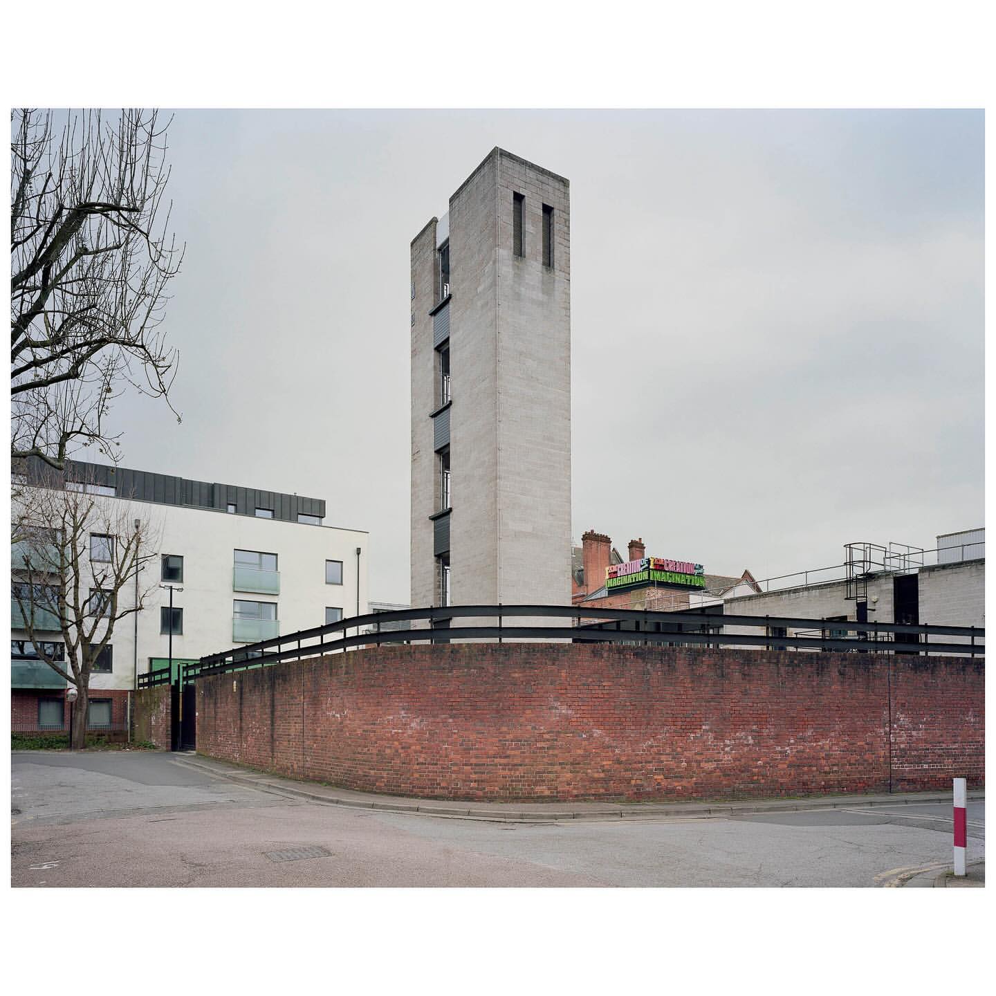 A tall concrete tower rises beside a red brick wall in an urban setting under a cloudy sky, framed by bare trees and adjacent buildings.