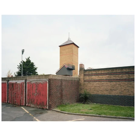 Urban scene features weathered red garage doors and a tall brick tower against a cloudy sky, creating an industrial yet austere landscape.