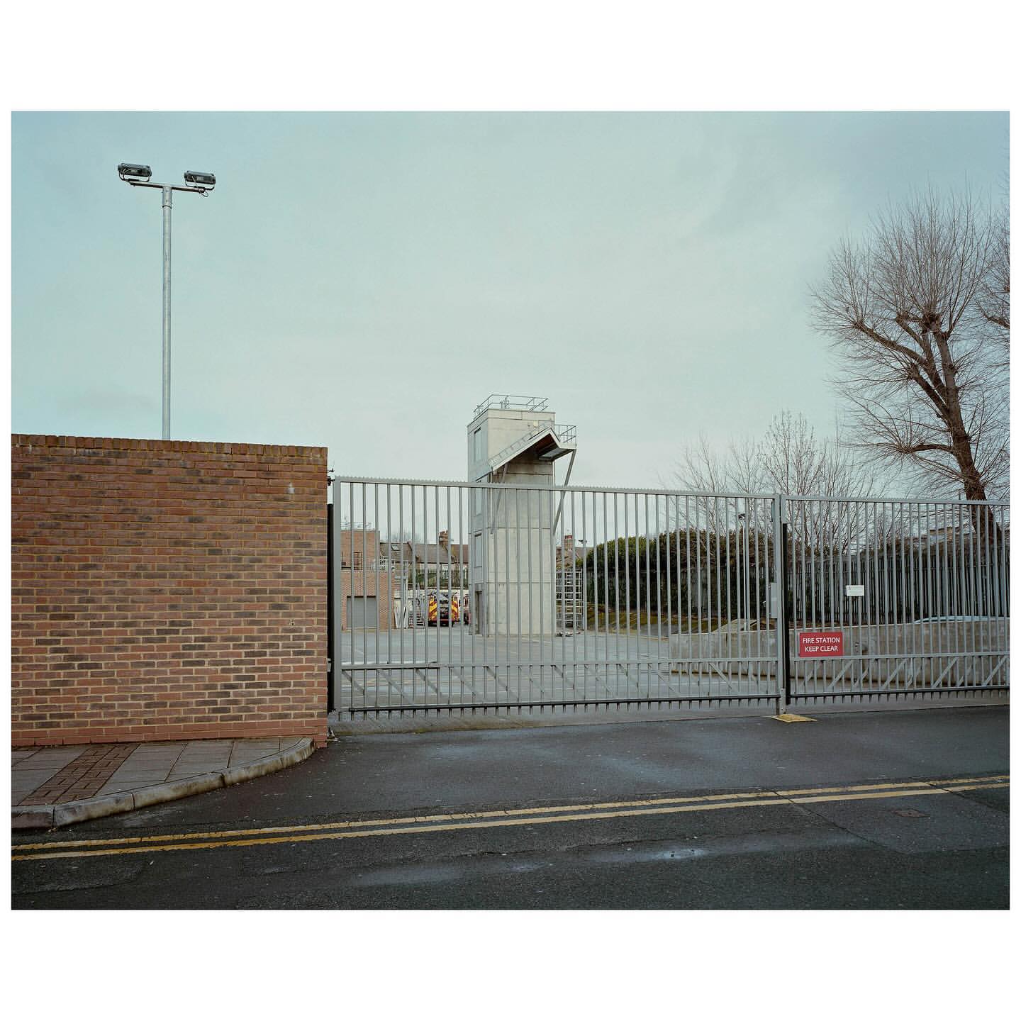 Exterior shot of a gated industrial complex featuring a tower and brick building under a cloudy sky, creating a muted, overcast ambiance.