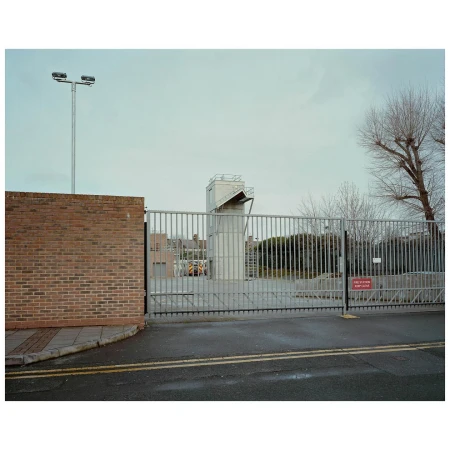 Exterior shot of a gated industrial complex featuring a tower and brick building under a cloudy sky, creating a muted, overcast ambiance.