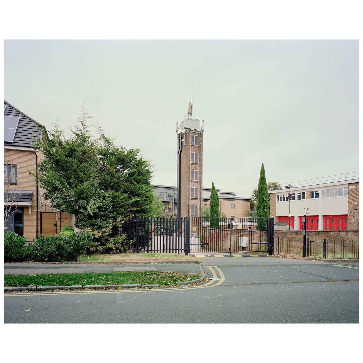 Exterior of a brick fire station with a tall brick tower against a cloudy sky, enclosed by a black metal fence and gate.