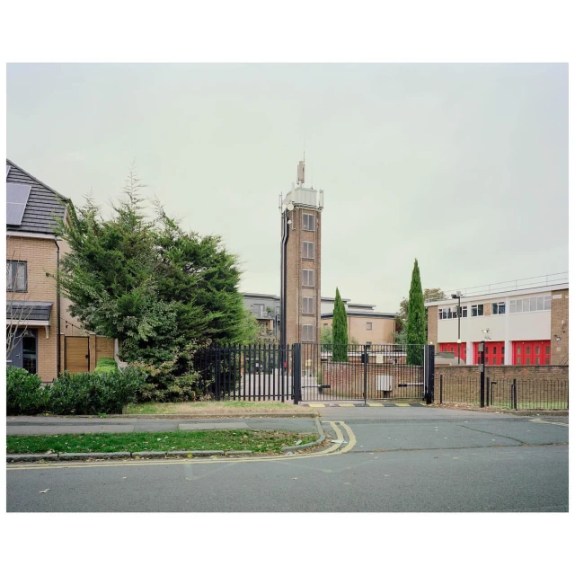 Exterior of a brick fire station with a tall brick tower against a cloudy sky, enclosed by a black metal fence and gate.