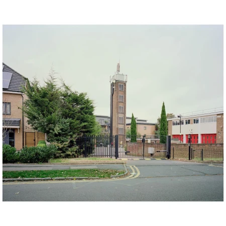 Exterior of a brick fire station with a tall brick tower against a cloudy sky, enclosed by a black metal fence and gate.