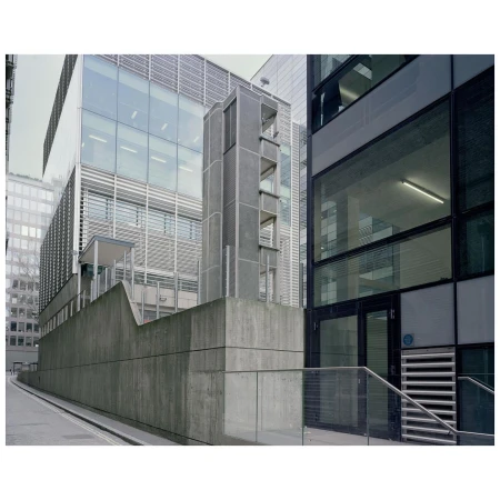 Modern commercial office exterior with concrete walls, glass facades, and a tall central tower structure against an overcast sky.
