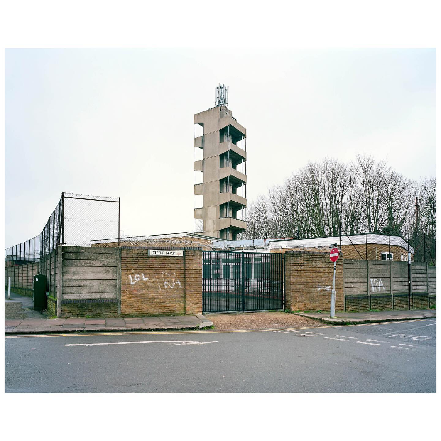 Exterior of concrete tower and brick building with graffiti, metal fence, and street sign on overcast day in urban setting.