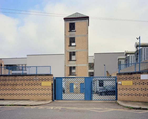 Exterior view of a brick and beige building with a tower, seen through a blue metal gate on an overcast day, urban landscape.