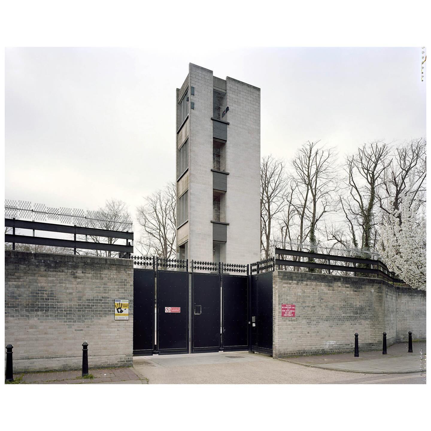 Exterior shot of a tall, concrete building with a gated entrance, surrounded by trees and a gray sky, creating an architectural image.