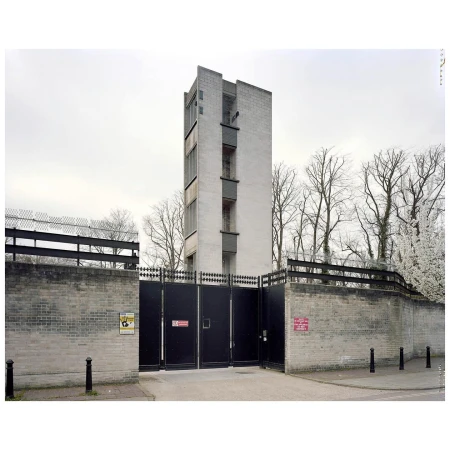 Exterior shot of a tall, concrete building with a gated entrance, surrounded by trees and a gray sky, creating an architectural image.