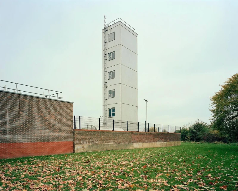 Tall white tower rises over a brick wall in an outdoor setting with fallen leaves on the ground, creating an architectural and natural landscape.