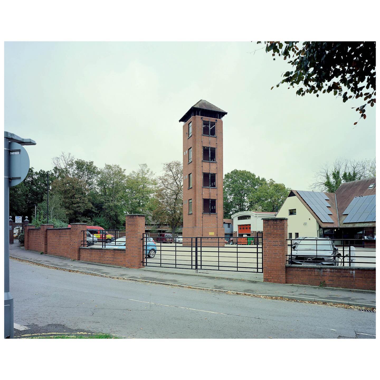 Brick tower in a parking lot surrounded by trees and buildings under an overcast sky with parked cars, walls and metal fencing.