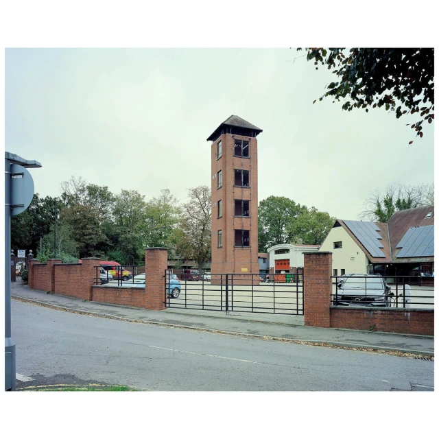 Brick tower in a parking lot surrounded by trees and buildings under an overcast sky with parked cars, walls and metal fencing.