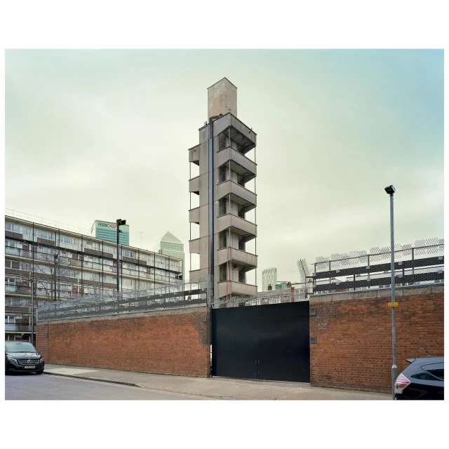 Exterior shot of a concrete tower with an open staircase, a brick wall, and modern buildings in the background under an overcast sky.