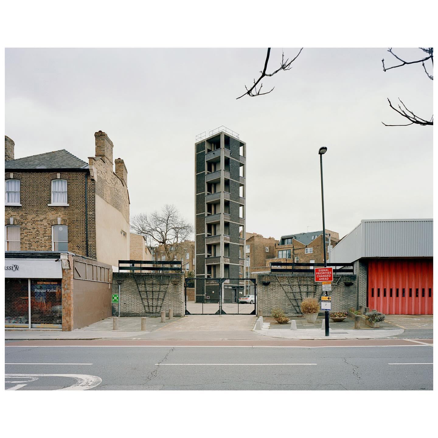 Urban scene featuring a narrow, modern concrete tower block between brick buildings, viewed from a street on an overcast day.