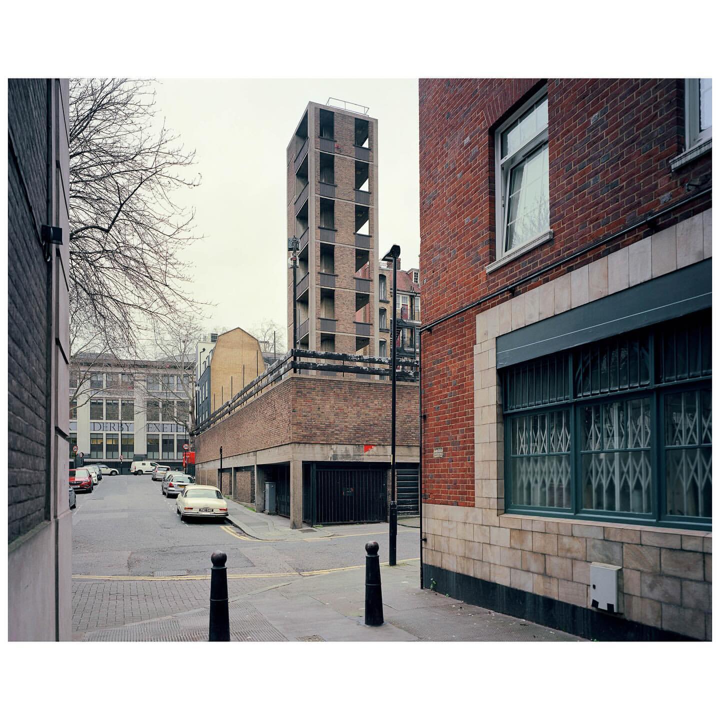 Urban street scene features a brick building with window bars and a tall, modern concrete tower in the background under an overcast sky.
