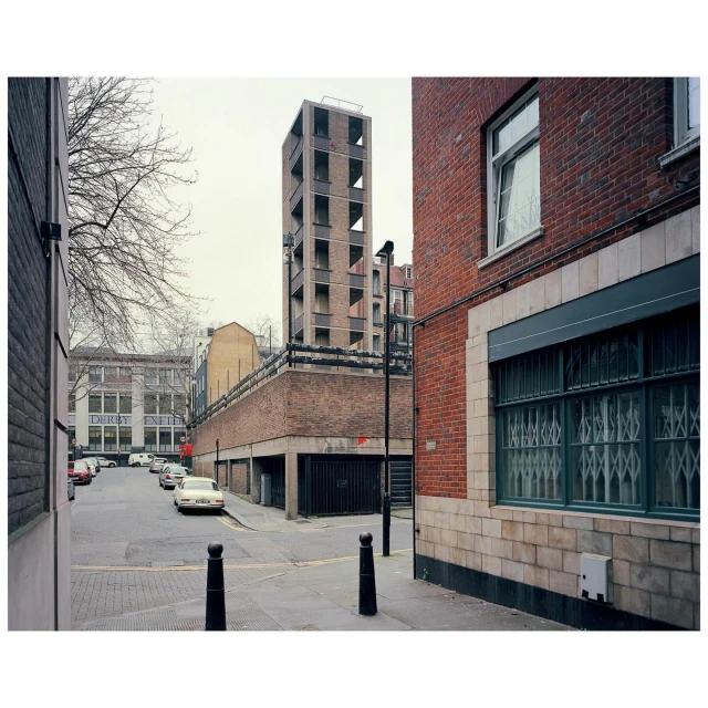 Urban street scene features a brick building with window bars and a tall, modern concrete tower in the background under an overcast sky.