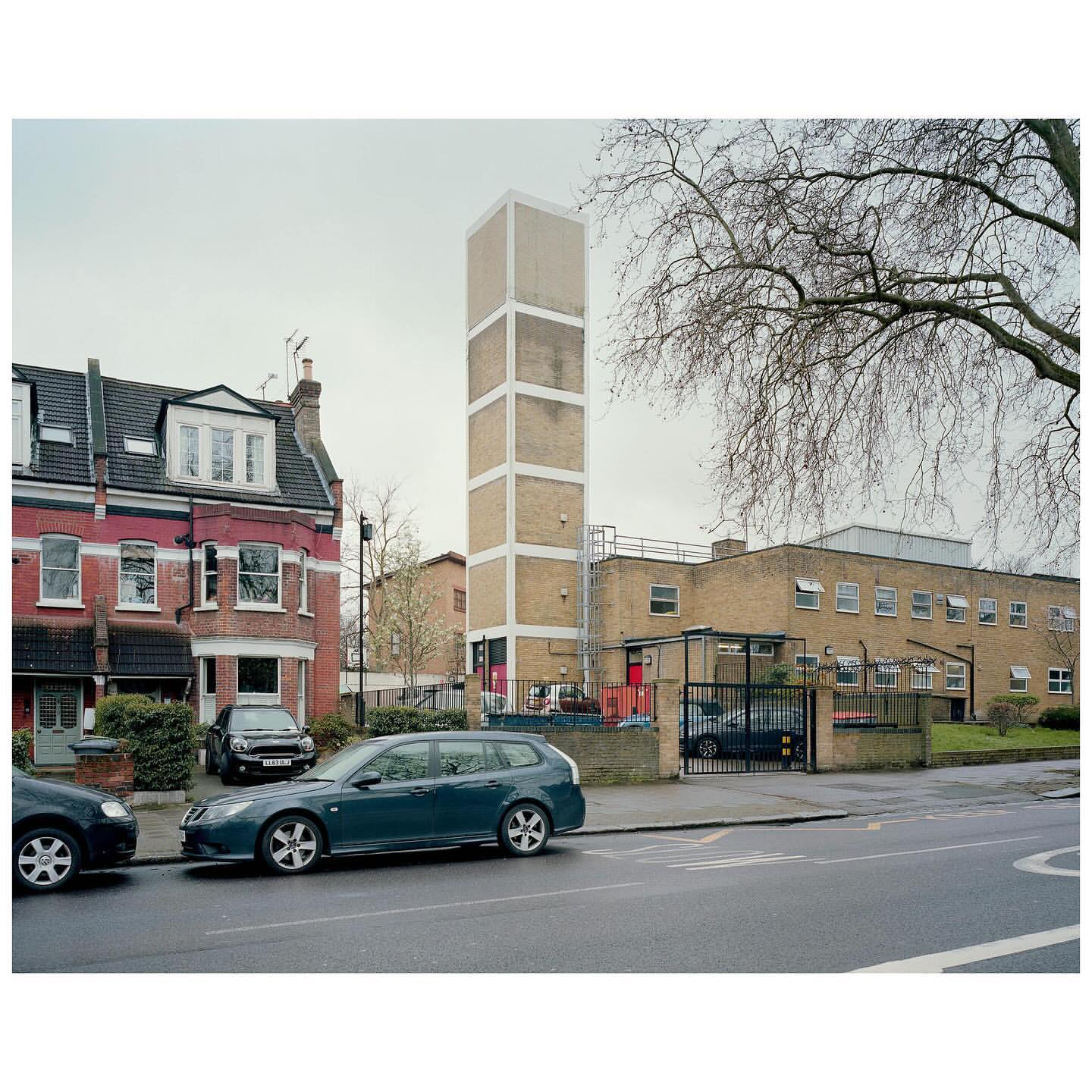 Urban scene featuring a tall, rectangular tower alongside a multi-story brick building and parked cars on a quiet, overcast day.