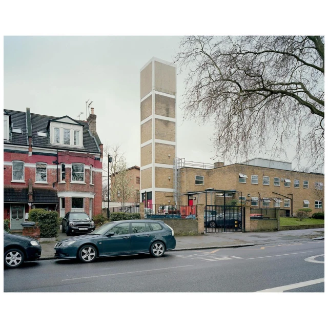 Urban scene featuring a tall, rectangular tower alongside a multi-story brick building and parked cars on a quiet, overcast day.