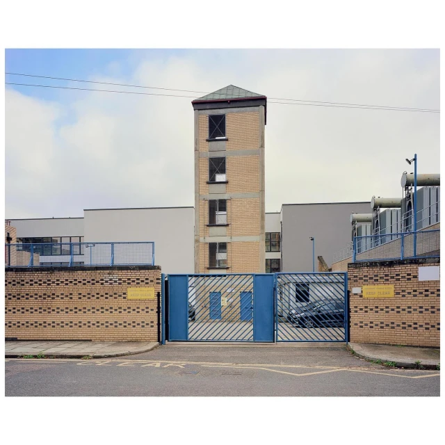 Exterior view of a tall brick tower building with a flat roof, situated behind a blue metal gate and brick walls, on a cloudy day.
