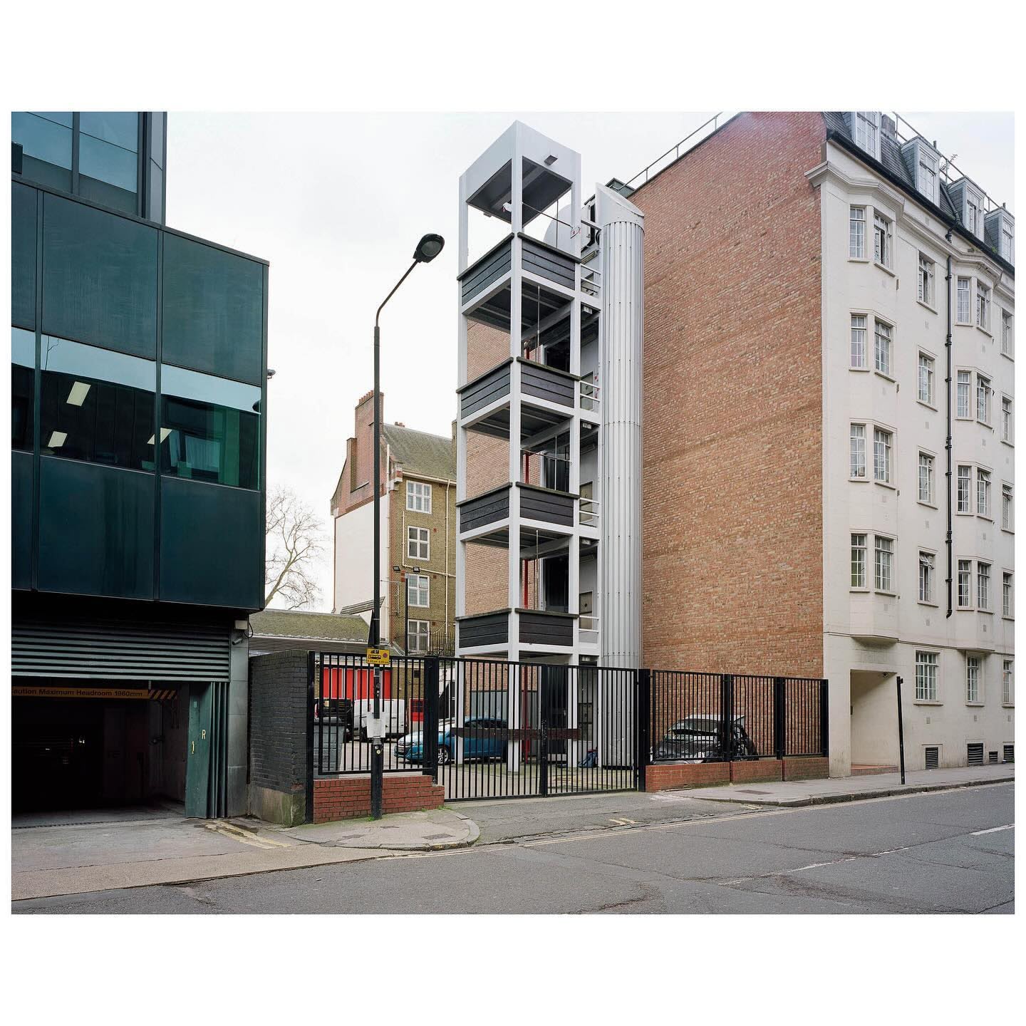 Exterior view of a modern building featuring an external staircase and a cylindrical structure, captured on a cloudy day in an urban environment.