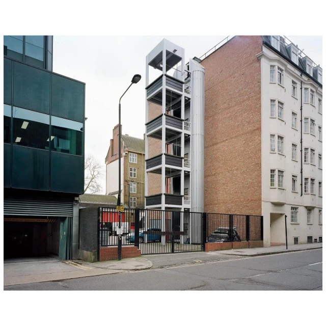 Exterior view of a modern building featuring an external staircase and a cylindrical structure, captured on a cloudy day in an urban environment.