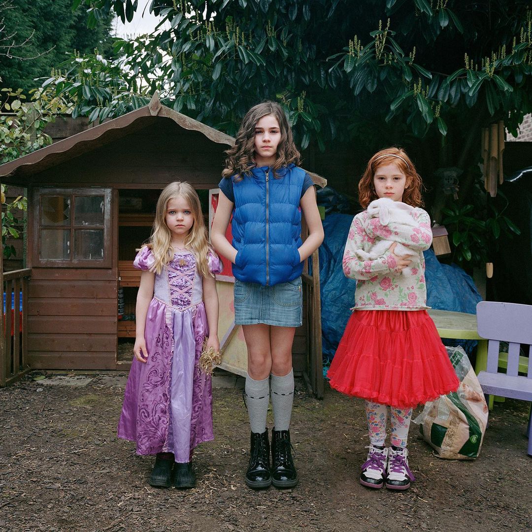 Three young girls stand outdoors in dresses in front of a playhouse and greenery, one holding a plush toy. Square composition.