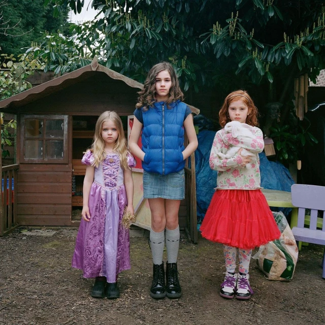 Three young girls stand outdoors in dresses in front of a playhouse and greenery, one holding a plush toy. Square composition.