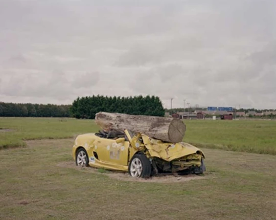 Damaged yellow convertible car with a large tree log on its hood, parked on a grassy field beneath a cloudy sky, with a tree line in the distance.