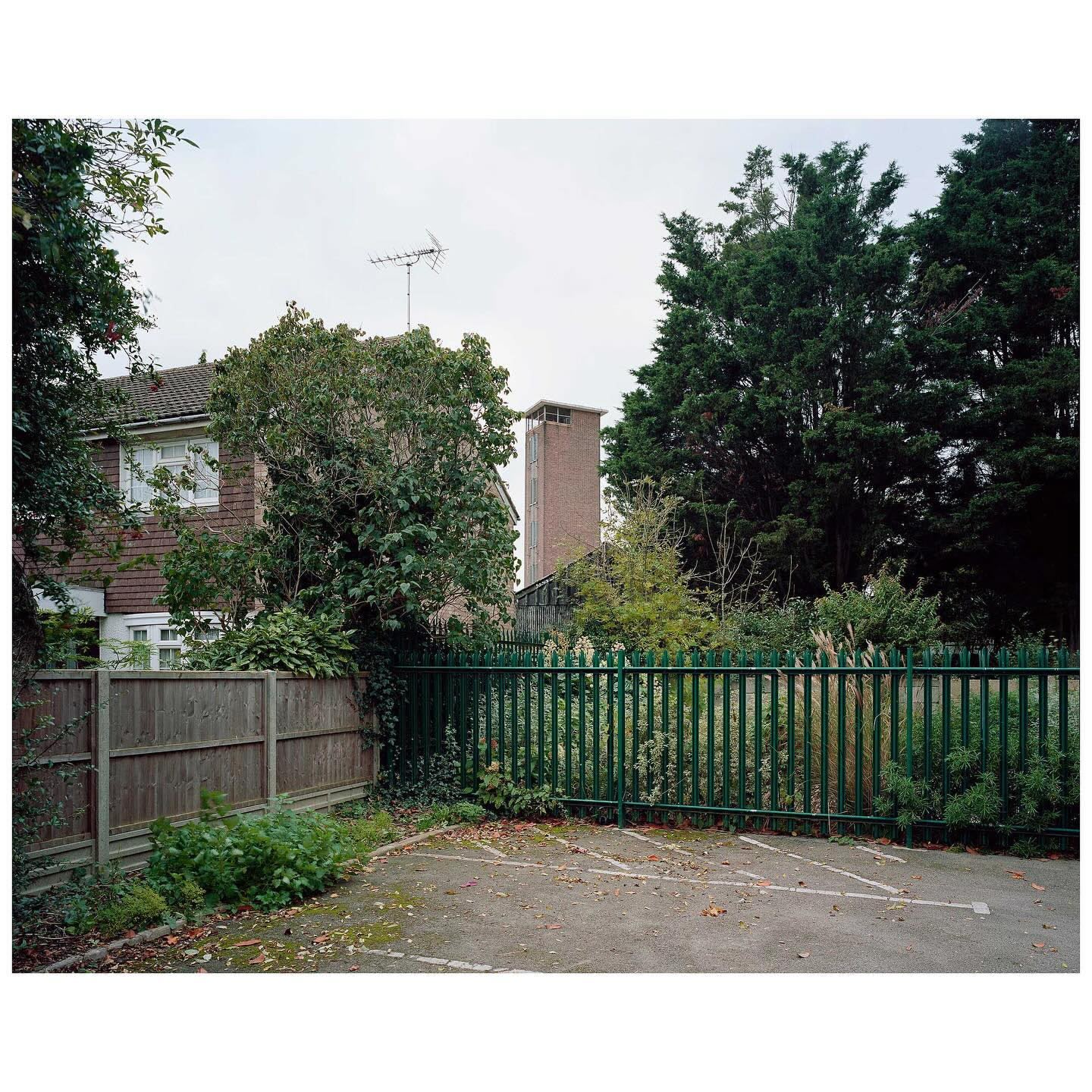 Exterior daytime shot of a two-story house behind a dark green metal fence with a paved parking area in the foreground.
