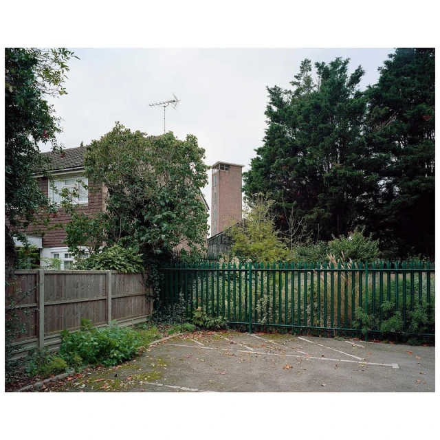 Exterior daytime shot of a two-story house behind a dark green metal fence with a paved parking area in the foreground.