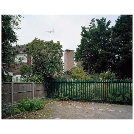 Exterior daytime shot of a two-story house behind a dark green metal fence with a paved parking area in the foreground.