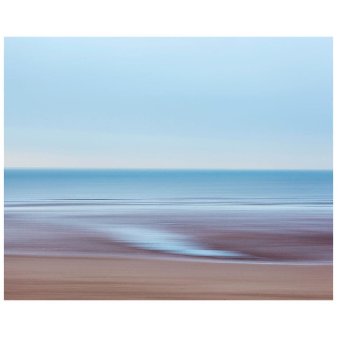 Blurred long exposure captures a tranquil seascape with a sandy beach, gentle waves, and a pastel blue sky, creating a serene scene.
