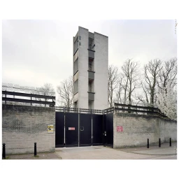 Tall concrete tower behind a brick wall with black metal gates and barbed wire, photographed by Paul Thompson.