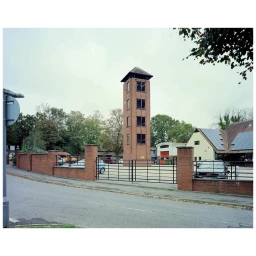 A tall brick training tower enclosed by a brick wall and metal gates behind an asphalt road.