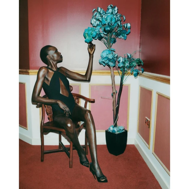 A dark-skinned woman in a black swimsuit sits in a wooden chair next to a decorative tree with blue flowers against a red wall.