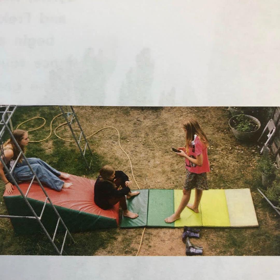Three children are outdoors in a grassy backyard, interacting on colorful play equipment with a garden hose in the background.