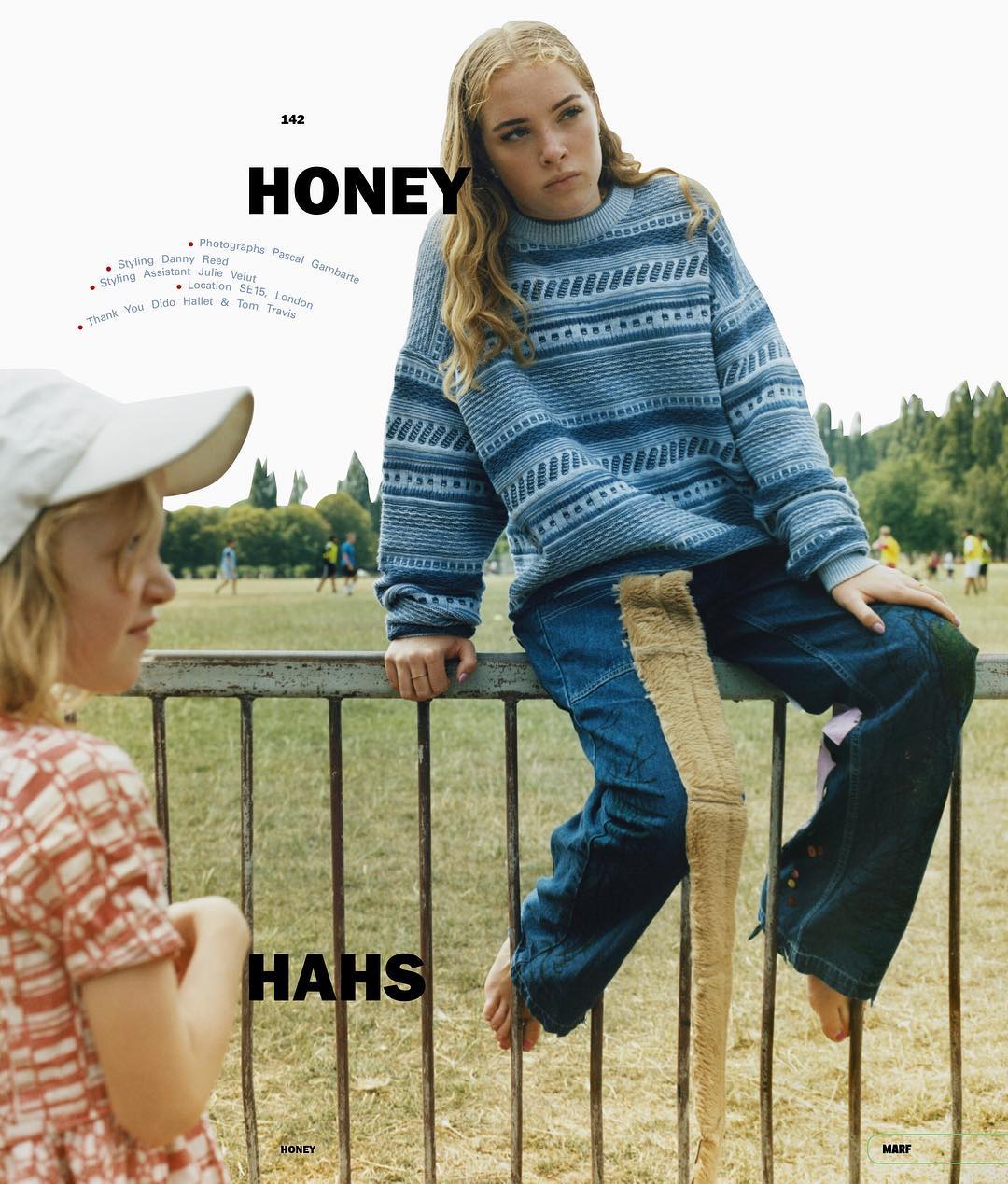 Two young girls outdoors; one in a patterned top looking to the left, and one in a blue patterned sweater sitting on a fence.