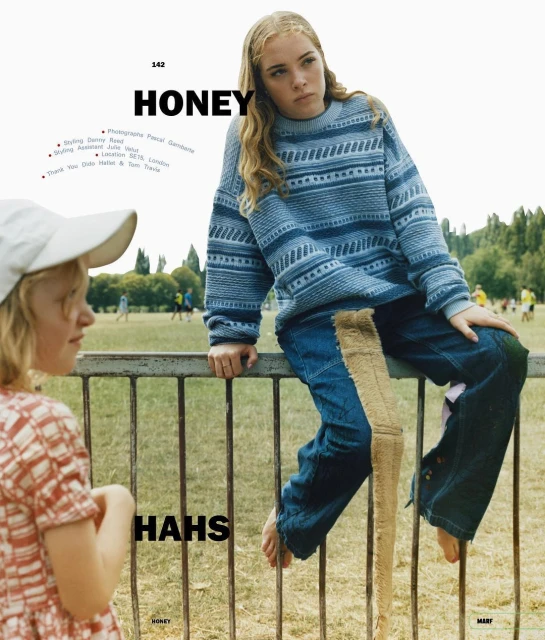 Two young girls outdoors; one in a patterned top looking to the left, and one in a blue patterned sweater sitting on a fence.