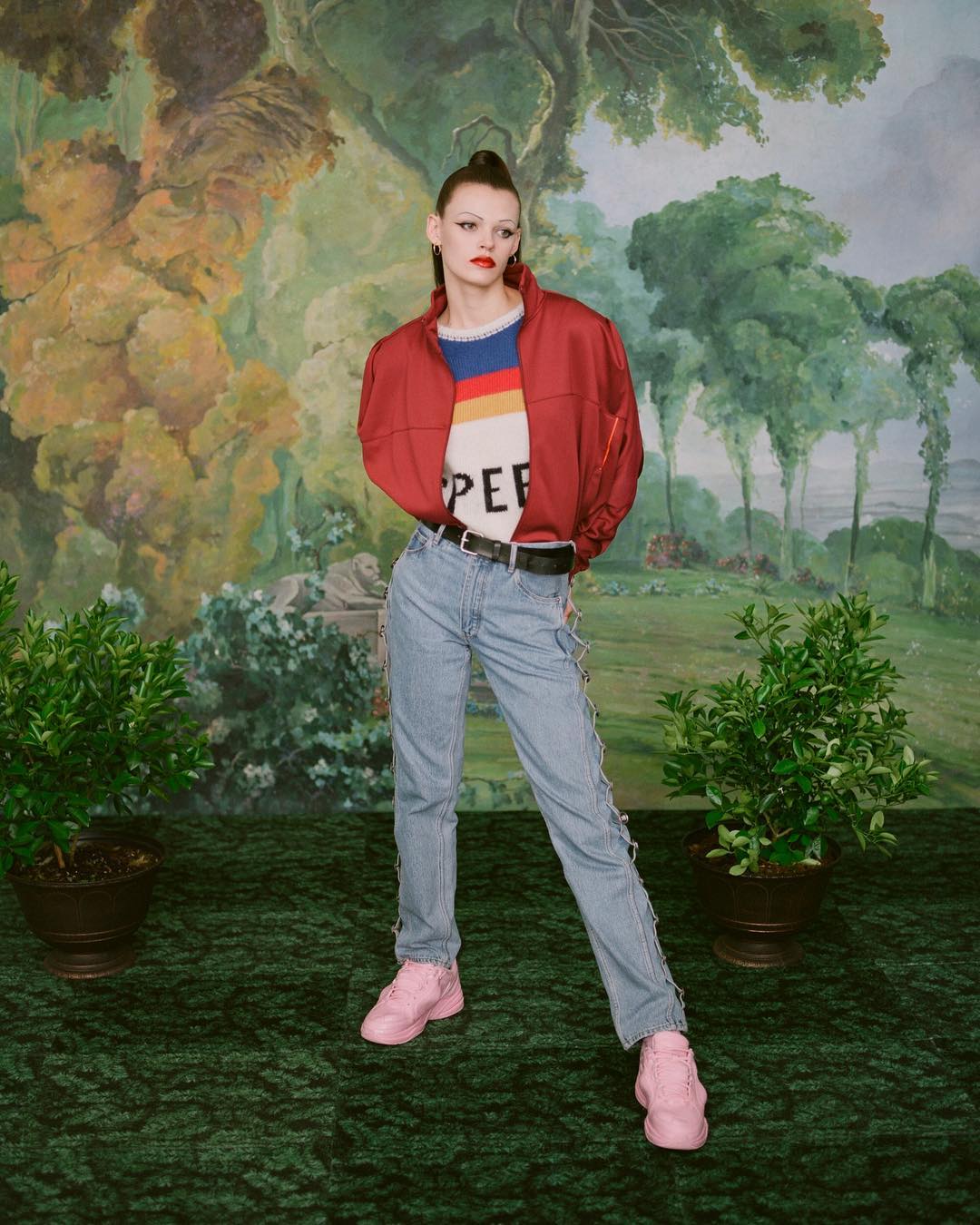 A young adult female fashion model poses in front of a painted backdrop wearing a red jacket, light blue jeans, and pink sneakers.
