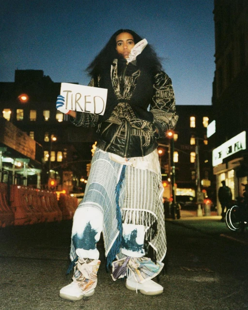 Woman in avant-garde fashion holds a 'TIRED' sign on a city street at dusk. Padded vest, patchwork pants, artistic sneakers, and dark hair.