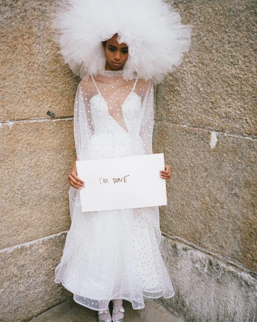 Woman in white tulle dress and large headpiece holds a sign in front of a stone wall, expressing a somber mood.