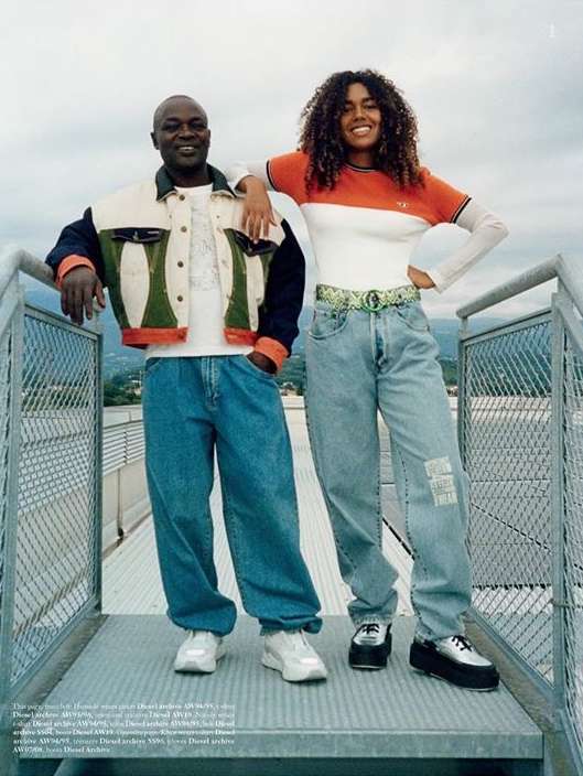 Two people, a man and a woman, are standing outdoors on a metal structure, wearing casual denim jeans and jackets, with a cityscape view.