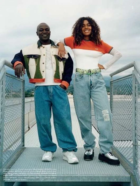 Two people, a man and a woman, are standing outdoors on a metal structure, wearing casual denim jeans and jackets, with a cityscape view.